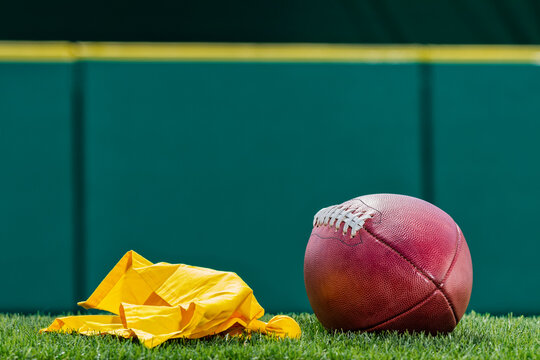 A low angle view of a yellow Penalty Flag thrown by a referee next to a pro American Football with green padded wall in the background. 