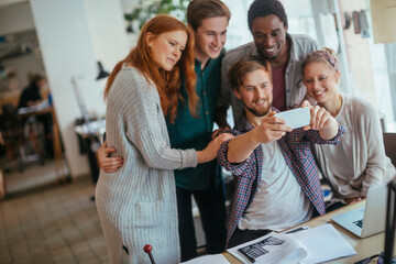 Young and diverse group of architects taking a selfie on a smartphone while working together in a startup company office