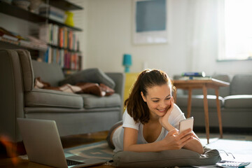 Obraz premium Young woman using her smartphone on the floor of her living room at home