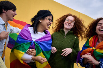 Group excited young diverse people laughing yellow wall background. LGBT community enjoying gay pride day outdoors. Liberal people celebrating street party. Rainbow flags and friends of generation z. - obrazy, fototapety, plakaty