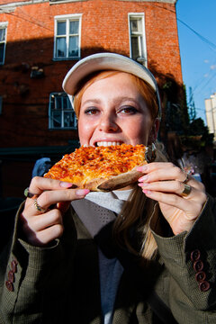 Junge schöne Frau mit einer Cappy isst ein größes stück Pizza auf der straße. im Hintergrund steht ein backteinhaus