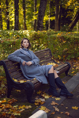 brunette woman in a gray coat and boots is sitting on a park bench in autumn among the trees. Orange foliage and a wooden bench during the day