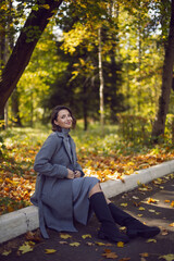 brunette woman in a gray coat and boots is sitting on a park curb in autumn among the trees. Orange foliage and a wooden bench during the day