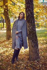 brunette woman in a gray coat and boots is standing by a tree in autumn among the trees. Orange foliage during the day