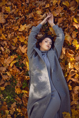 brunette woman in a gray coat and boots it lies on the foliage in autumn. Orange foliage during the day
