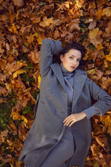 brunette woman in a gray coat and boots it lies on the foliage in autumn. Orange foliage during the day
