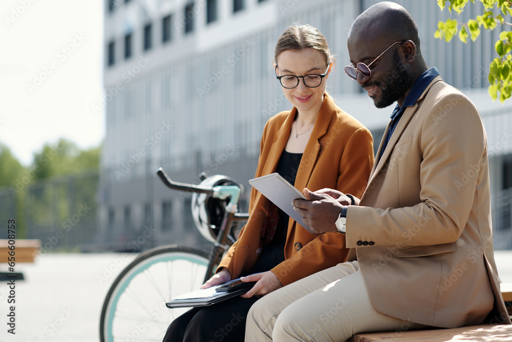 Poster Young businesswoman and her male colleague looking at tablet screen while sitting in front of camera and discussing points of project
