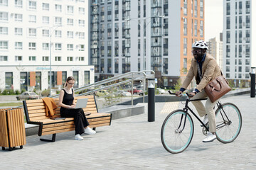 Happy young businessman in formalwear and safety helmet moving by businesswoman with laptop while riding bicycle in urban environment