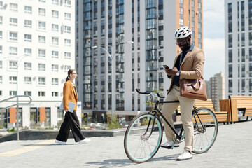 Young smiling male entrepreneur in safety helmet sitting on bicycle and scrolling through online information in mobile phone in the street