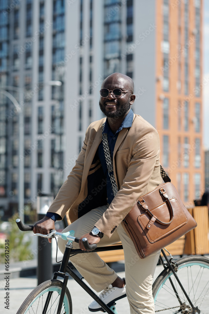 Poster young successful man in quiet luxury attire sitting on bicycle in front of camera and looking at you