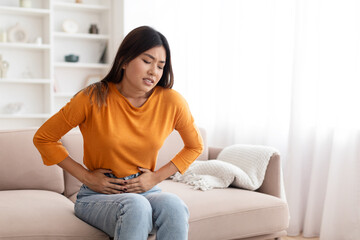 Unhappy sick young asian woman sitting on couch, touching belly - obrazy, fototapety, plakaty