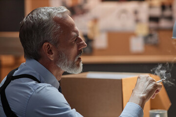 Side view portrait of handsome senior detective smoking cigarette in office