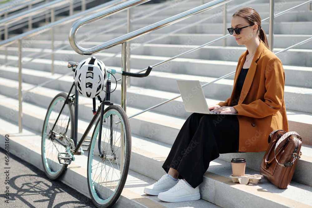 Poster young serious female solopreneur with laptop sitting on staircase in front of camera and organizing 