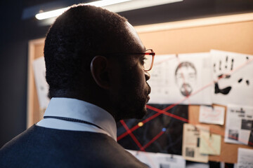 Dramatic back view of African American male detective looking at evidence board investigating case, copy space