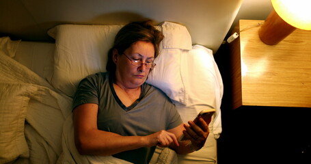 Older woman using smartphone device in bed looking at phone screen before sleep