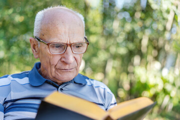 Concentrated senior man in eyewear seated in garden chair reads old book with yellow pages close up