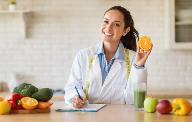 Happy female nutrition adviser holding orange fruit, making meal plan for client and smiling at...