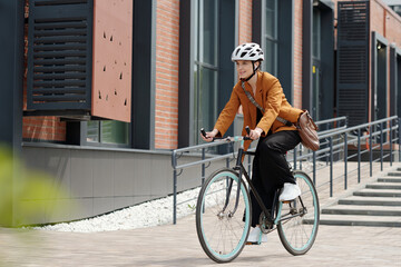 Young woman in formalwear and safety helmet sitting on bicycle and riding home from work while moving along modern building - obrazy, fototapety, plakaty