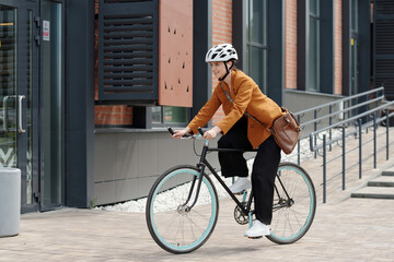 Side view of young businesswoman in quiet luxury attire and protective helmet riding bicycle along...