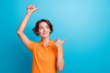 Photo of charming thoughtful lady wear orange t-shirt pointing two thumbs looking empty space isolated blue color background