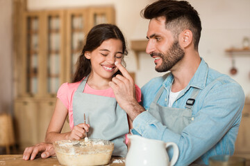 Loving dad and his daughter baking in kitchen, having fun, father playfully touching girl's nose with flour and smiling