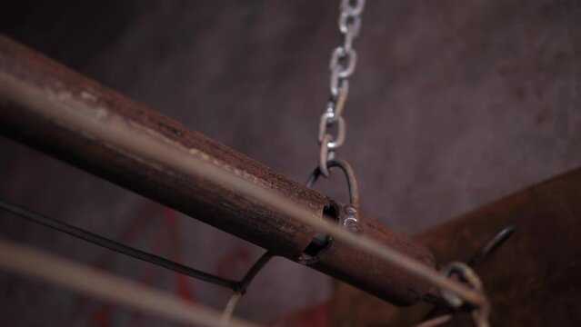 A close-up of a metal structure made of pipe and rusty iron rods hanging on chains in a dark creepy room.