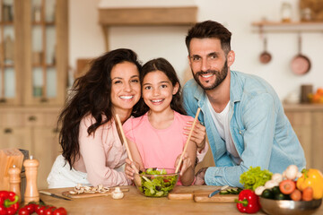 Happy european family cooking together and smiling to camera, parents embrace daughter while girl mixing salad