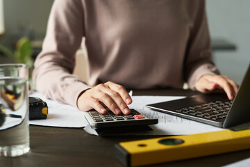 Focus on hand of young female architect and accountant using calculator and laptop while sitting by...