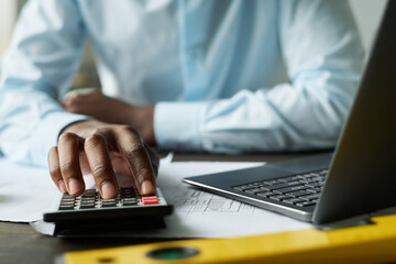 Hand of young African American male accountant using calculator by workplace while counting expenses on new engineering project - obrazy, fototapety, plakaty