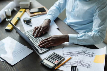 Close-up of young modern male architect keeping hands on laptop keyboard while sitting by workplace with blueprints and documents