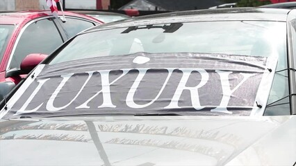 luxury word text sign banner flag on front of black car vehicle windshield at dealership outside exterior on bright day with confetti decoration waving in car hood reflection and flag blowing behind