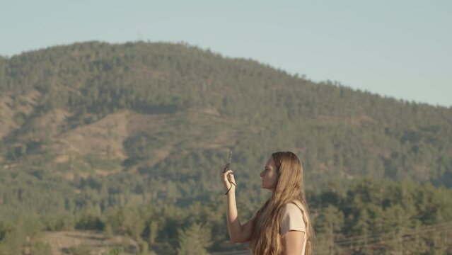 A young woman with long hair captures beautiful mountain views with trees on her mobile phone.