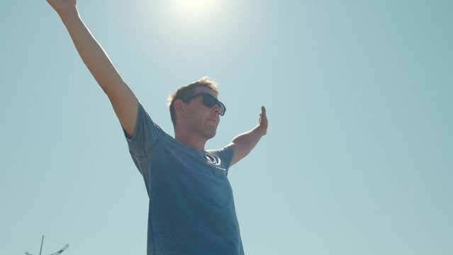 Happy Relaxed Man In Sunglasses Breathing Fresh Air Raising Arms Over Blue Sky With Sun. Dreaming, Freedom And Summer Travelin