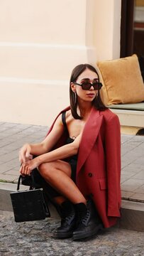 a beautiful business lady girl sits on a bench on the street in the foreground a red car drives by. vertical video. a girl in a jacket and a black bag in sunglasses is posing