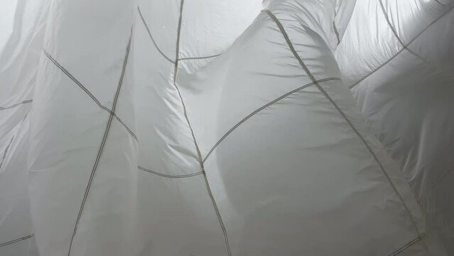 A huge parachute suspended from the ceiling in the pavilion sways in the wind, waves of white thin fabric, light shines through the fabric