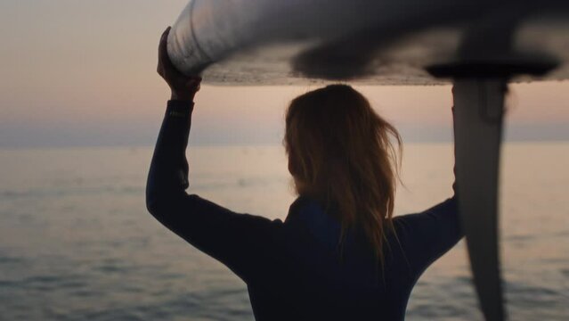Senior woman on the beach of the sea in a wetsuit goes into the water. Granny the female is surfing on a paddle board at sunset at dusk. Active old age on the ocean. Holds the board above his head