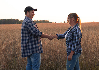 Naklejka na ściany i meble A beautiful couple of farmers at sunset inspects a field with wheat before selling grain Make notes on the tablet. Farmers smile and shake hands at sunset in the field. Agricultural business concept.