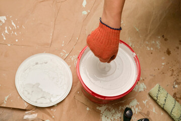 A worker in work gloves mixes white paint in a bucket and is going to paint the walls at home, preparing the paint before whitewashing © Мар'ян Філь