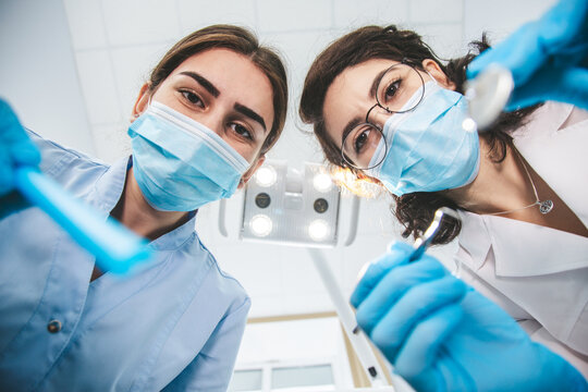Two female dentists examining patient's mouth from above using medical equipment, view from patient's point