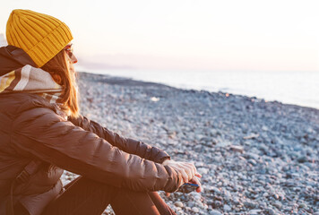 Portrait of a young woman in sunglasses, yellow knitted hat, scarf and with a backpack rests on a pebble beach by the sea in the evening and looks into the distance at the sunset in winter