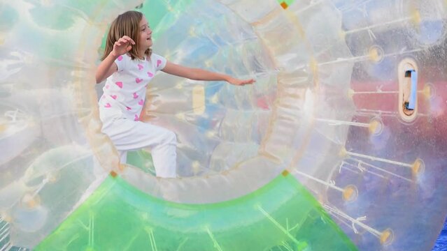 Cute cheerful eight year old blond girl running inside a light green aqua zorbing cylinder in an amusement park