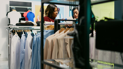 Young people talking about fashion collection in store, customer and employee looking at trendy new clothes in boutique. Two women analyzing retail shop merchandise at shopping center.