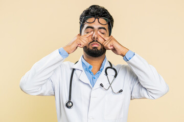 Exhausted tired Indian young doctor cardiologist man takes off glasses, feels eyes pain, being...