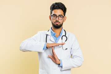 Tired serious upset Indian young doctor cardiologist man showing time out gesture, limit or stop sign, take a break, relax, rest, help. Arabian apothecary pharmacy guy isolated on beige background - obrazy, fototapety, plakaty