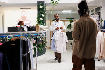 Clothes retail store african american man worker carrying shirts for customer to try. Buyer...