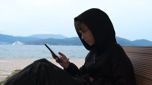 Depressed teenage girl sitting alone in the park by the bay with smart phone. Upset adolescent teenager outcast wearing hood feeling worried looks at the phone. Social media bullying concept.