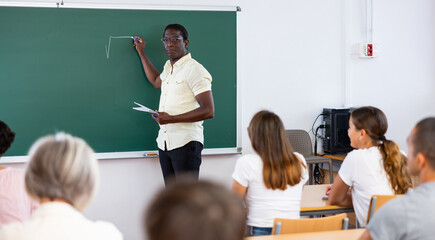 Portrait of confident african american professor giving lecture to group of students, standing near chalkboard and writing formulas