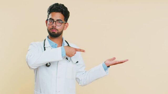 Indian young doctor cardiologist man show thumbs up and pointing right empty place, advertising area for commercial discount, copy space. Arabian apothecary pharmacy guy isolated on beige background