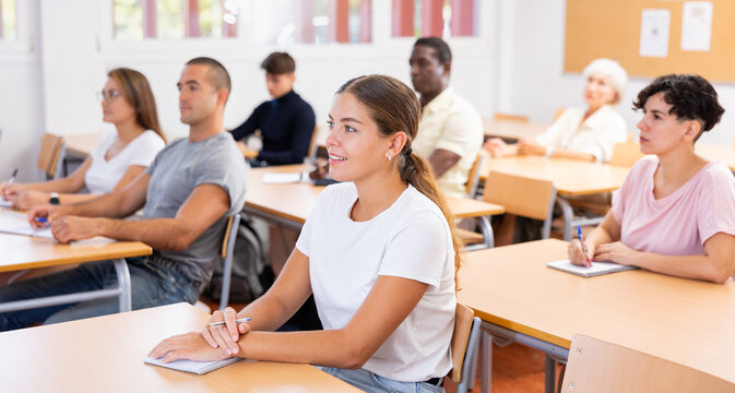 Smiling interested young girl attending courses of vocational retraining program for adults, listening to lecture and taking notes while sitting at desk in auditorium..