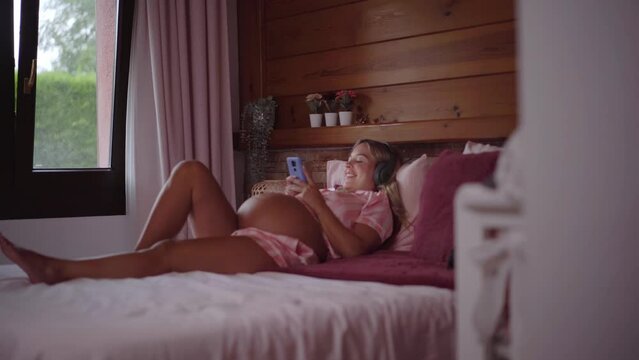 Young blonde pregnant lying on bed looking at mobile and listening to music headphones. Cheerful mom in third trimester belly using phone. Caucasian happy woman enjoying pre maternity at home.